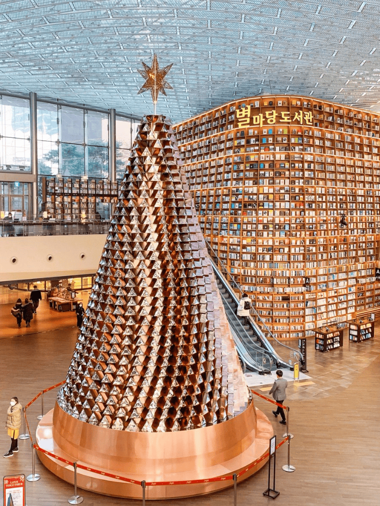 Shiny copper Christmas tree sculpture inside a modern bookstore with wooden shelves.