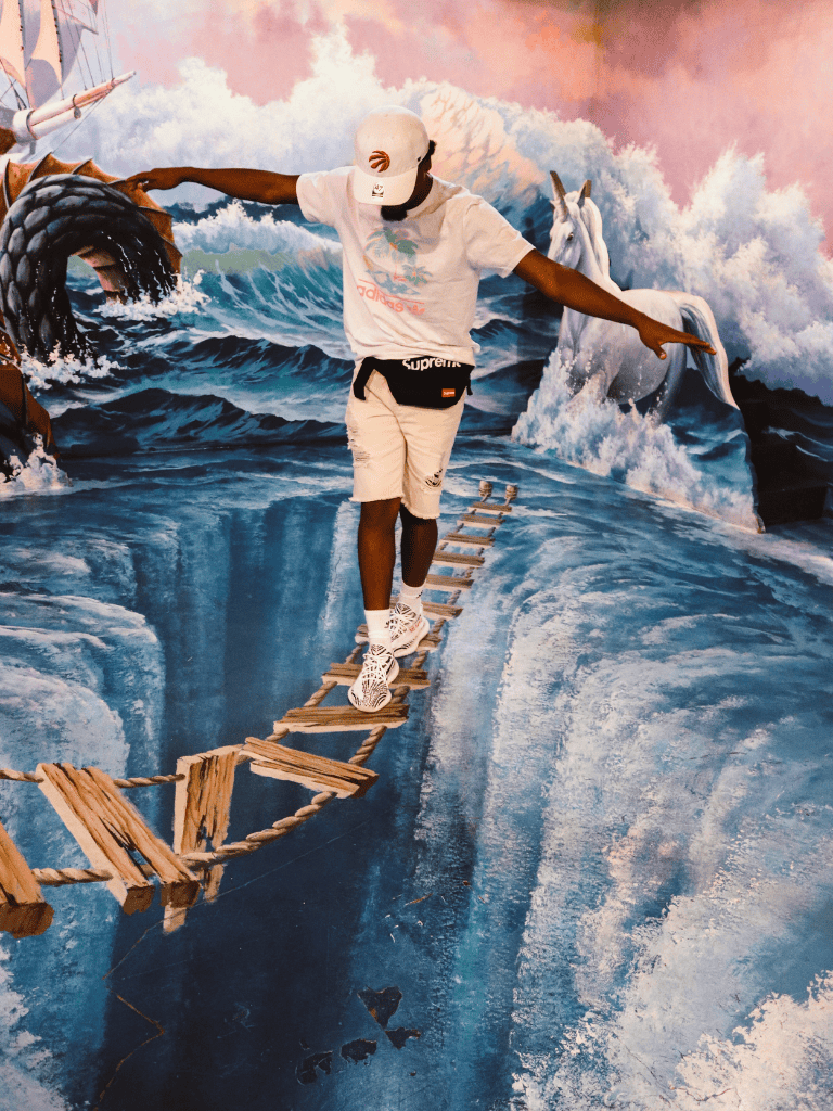 Brightly painted ocean scene with a person walking on a wooden rope bridge and a unicorn in the background.