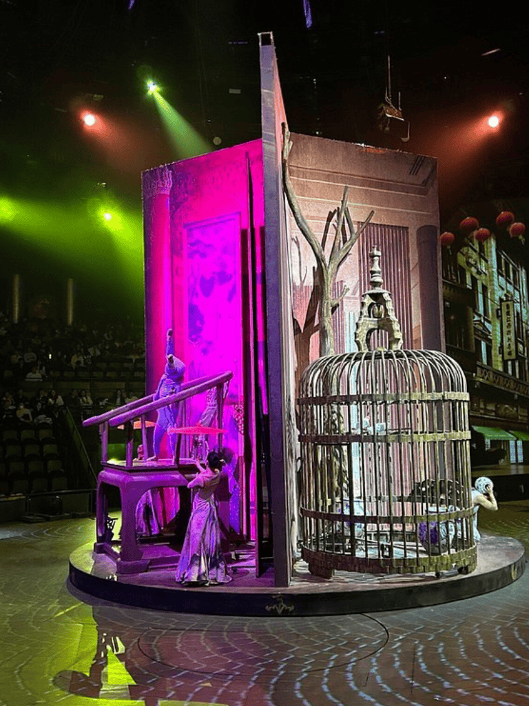A colorful theatrical stage set with elegant costumes, cage props, and vibrant lighting effects for an immersive performance.