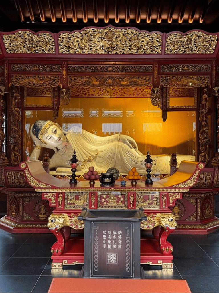 Reclining Buddha statue in ornate temple with intricate wood carvings and offerings.