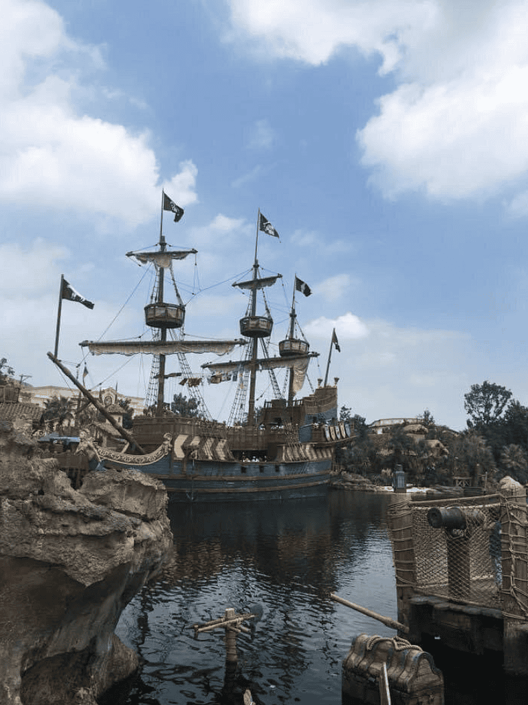 Pirate ship in a themed amusement park set against a cloudy sky with water and rocky surroundings.