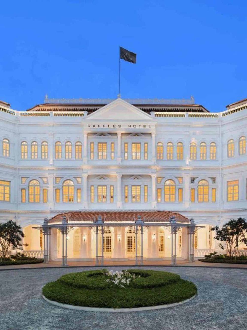 Elegant white hotel building at dusk with illuminated windows and the "Raffles Hotel" sign, showcasing luxury accommodation.