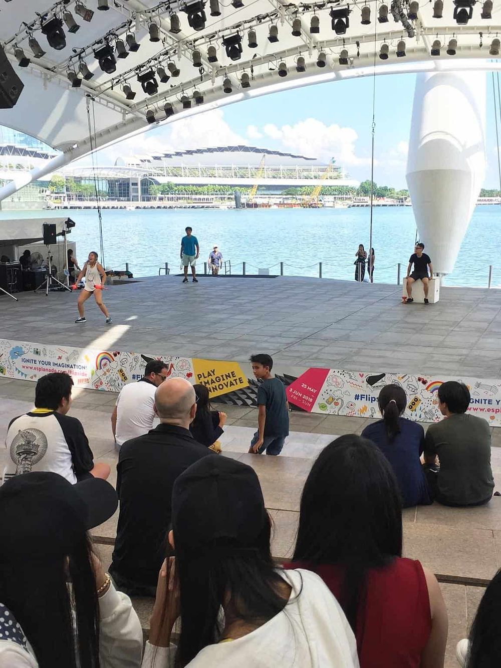 1. Modern outdoor stage at waterfront with performers and audience, Singapore skyline in background.