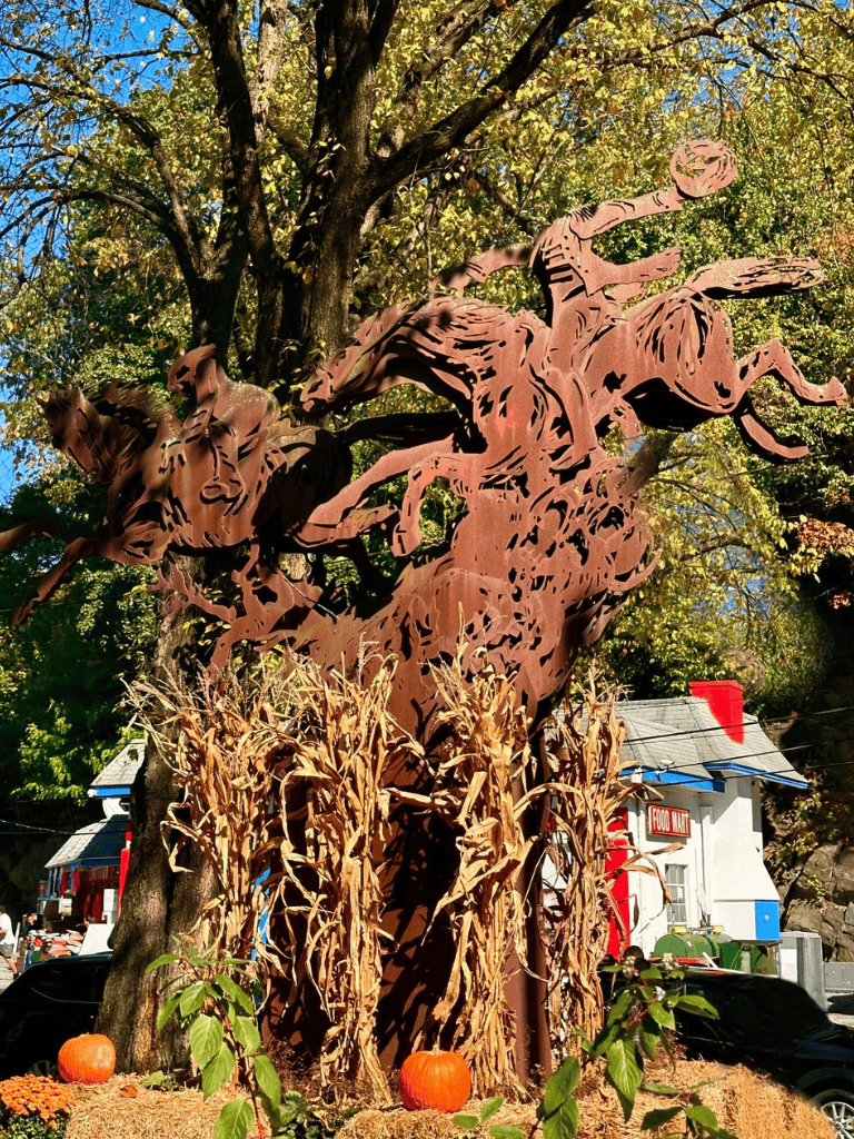 Rust-colored metal sculpture of a cowboy riding a bucking horse amidst fall scenery with pumpkins.