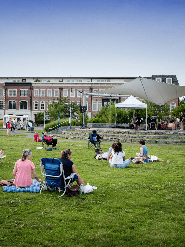 Relaxing outdoor concert in an urban park with people sitting on grass, enjoying live music under tents.