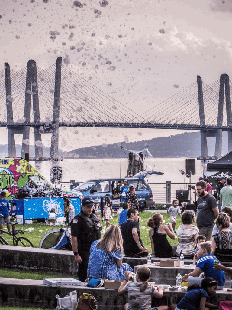 Fun family outdoor event at waterfront with kids, music, food, games, and entertainment, under a bridge.