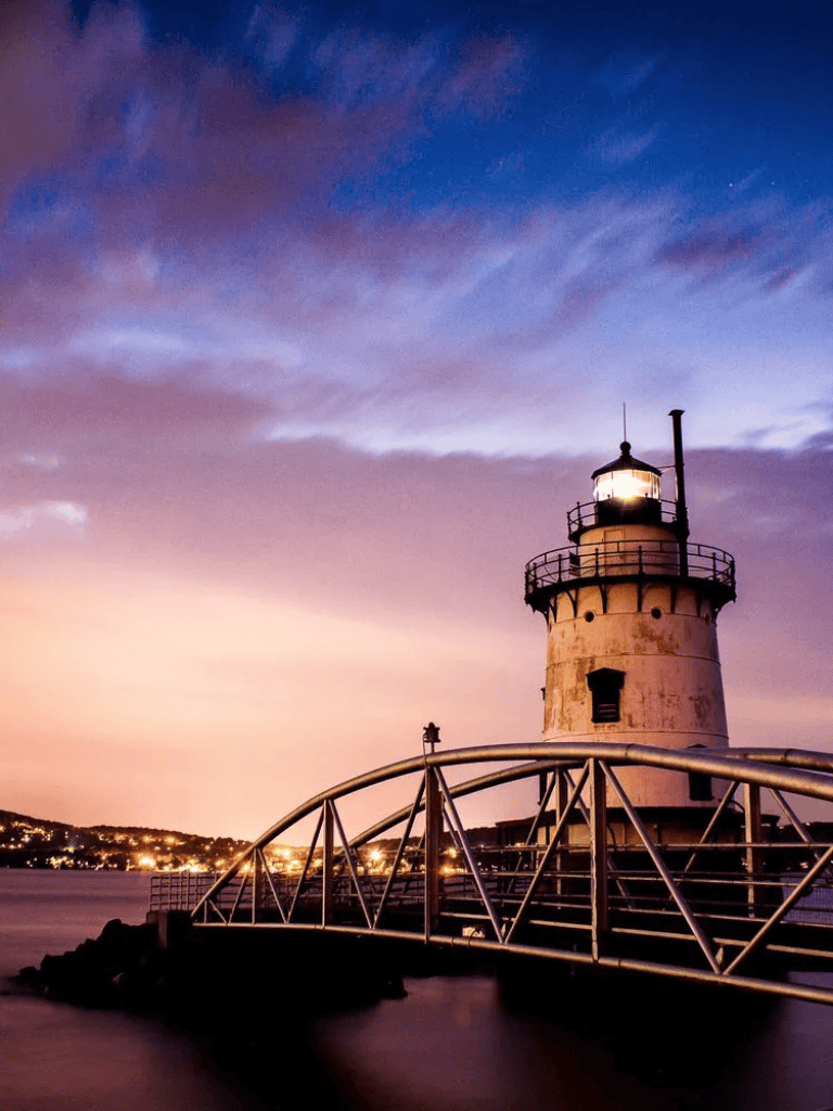 Lighthouse at sunset over the water with a bridge in the foreground, scenic coastal navigation.