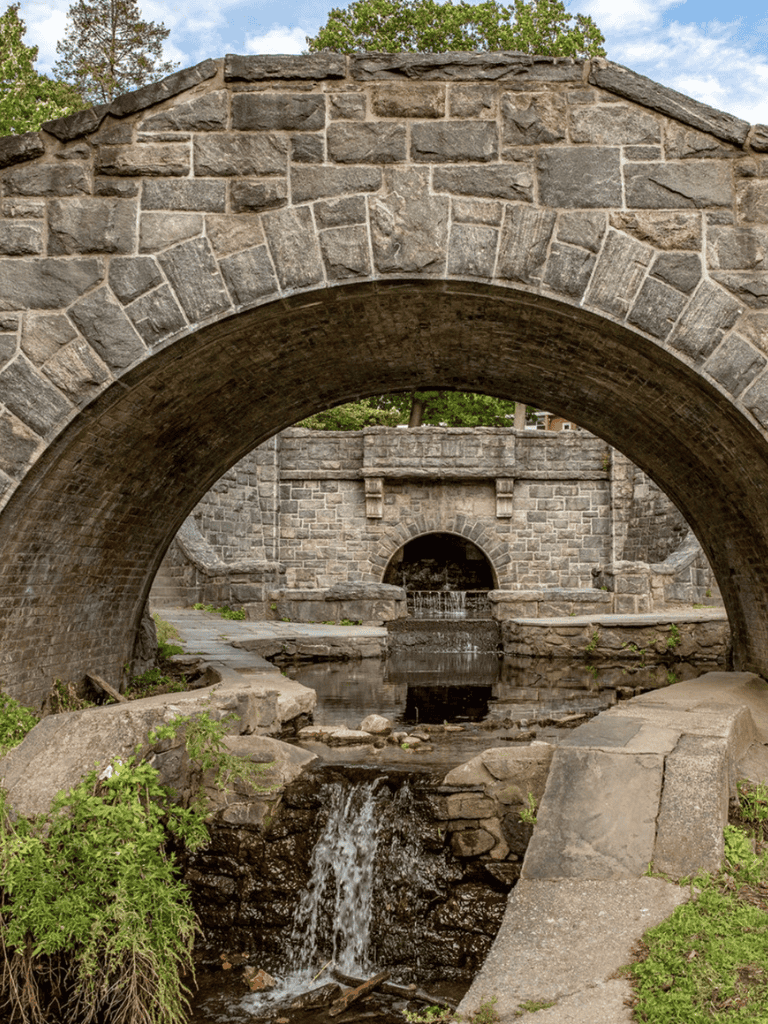 Old stone bridge with a water stream underneath, scenic historic structure in a park setting.