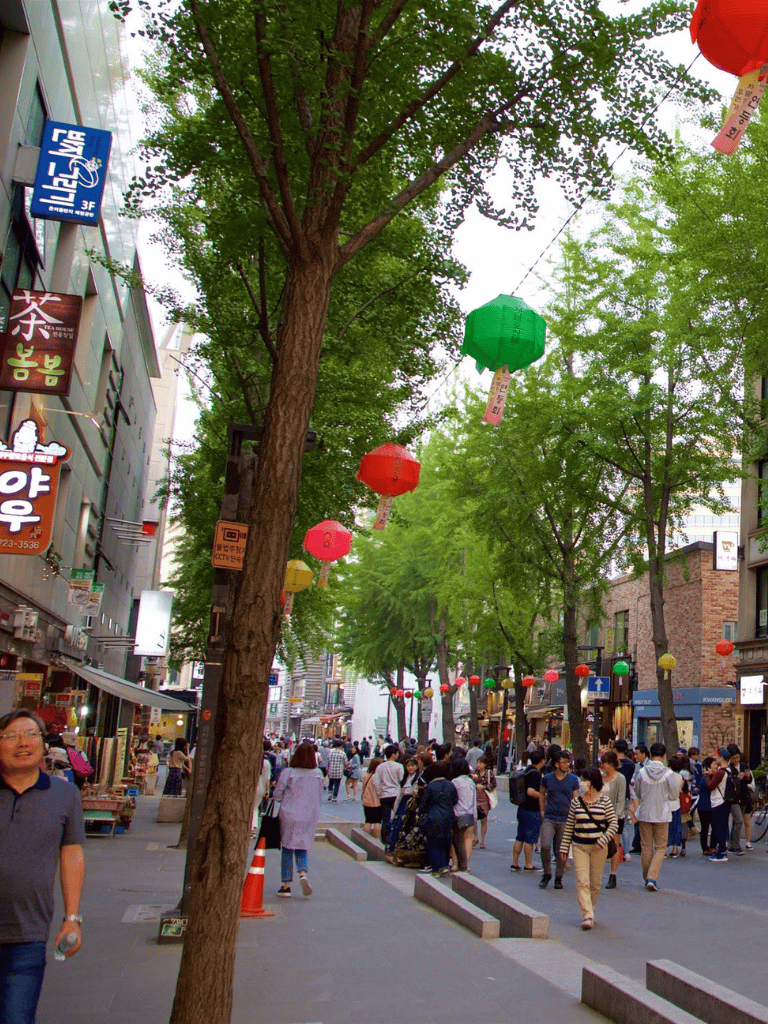 Colorful lanterns hanging over a busy city street with pedestrians, shops, and green trees.