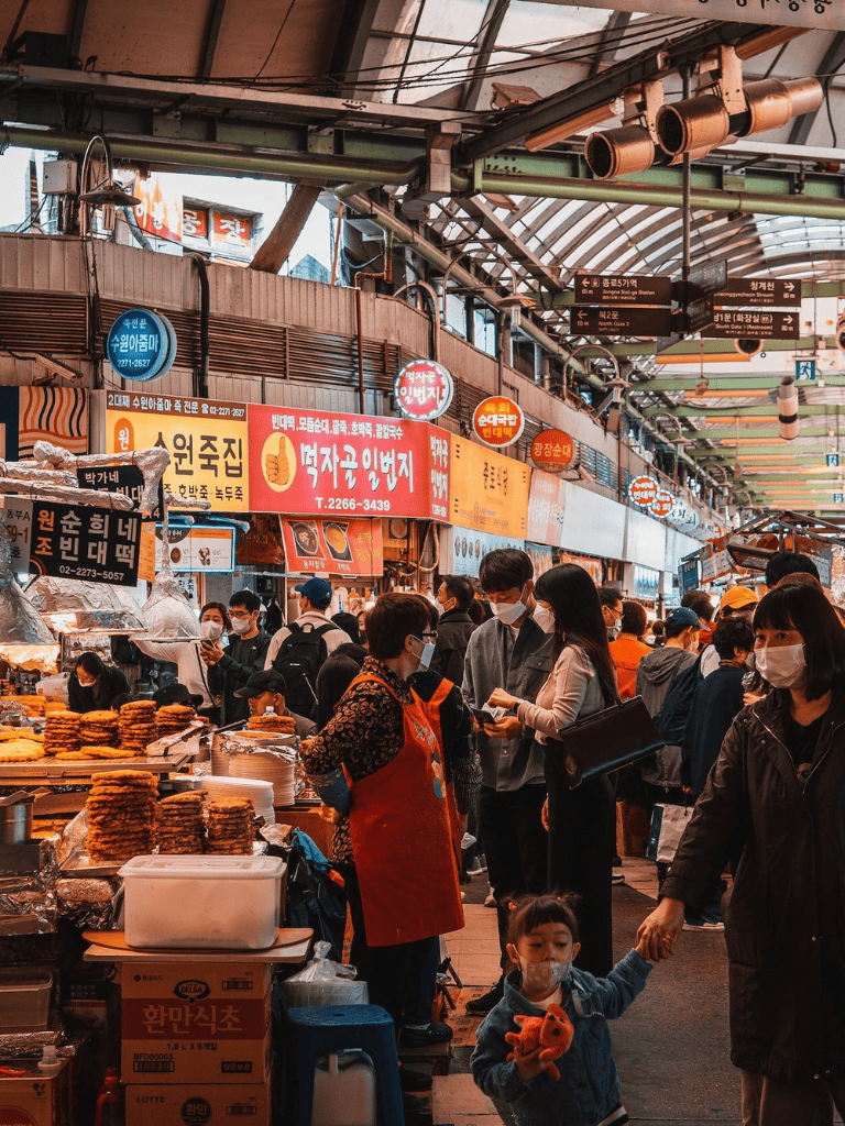 Busy indoor market with food stalls and shoppers, vibrant atmosphere, Korean signage, and diverse crowd.
