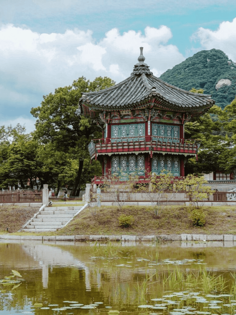 Traditional Korean pavilion near a pond with lush greenery and mountain backdrop.