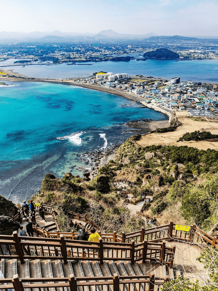 Vibrant coastal cityscape with stairway leading down to a blue bay, scenic views, and surrounding mountains.