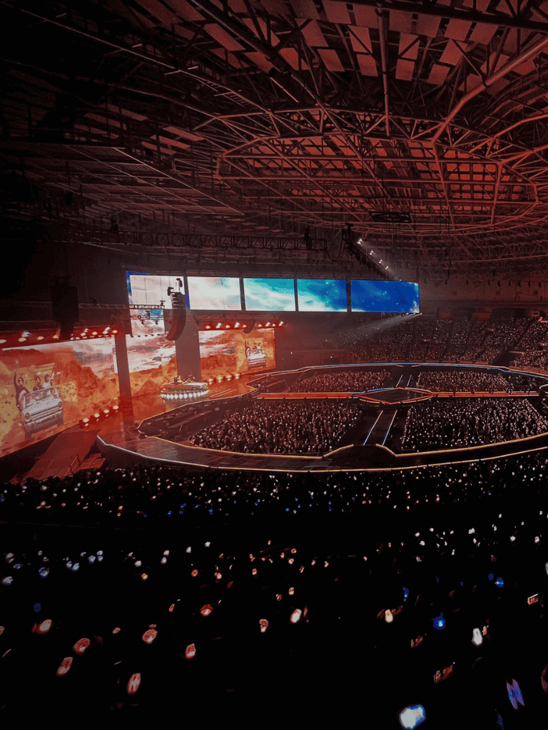 Brightly lit indoor concert with large LED screens and a full audience in a modern stadium.