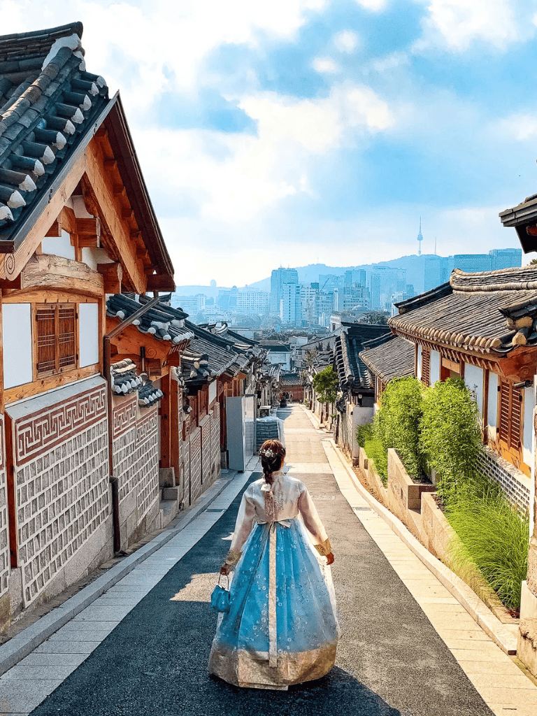 Traditional Korean hanbok on a woman walking through Bukchon Hanok Village, Seoul, with city skyline in background.