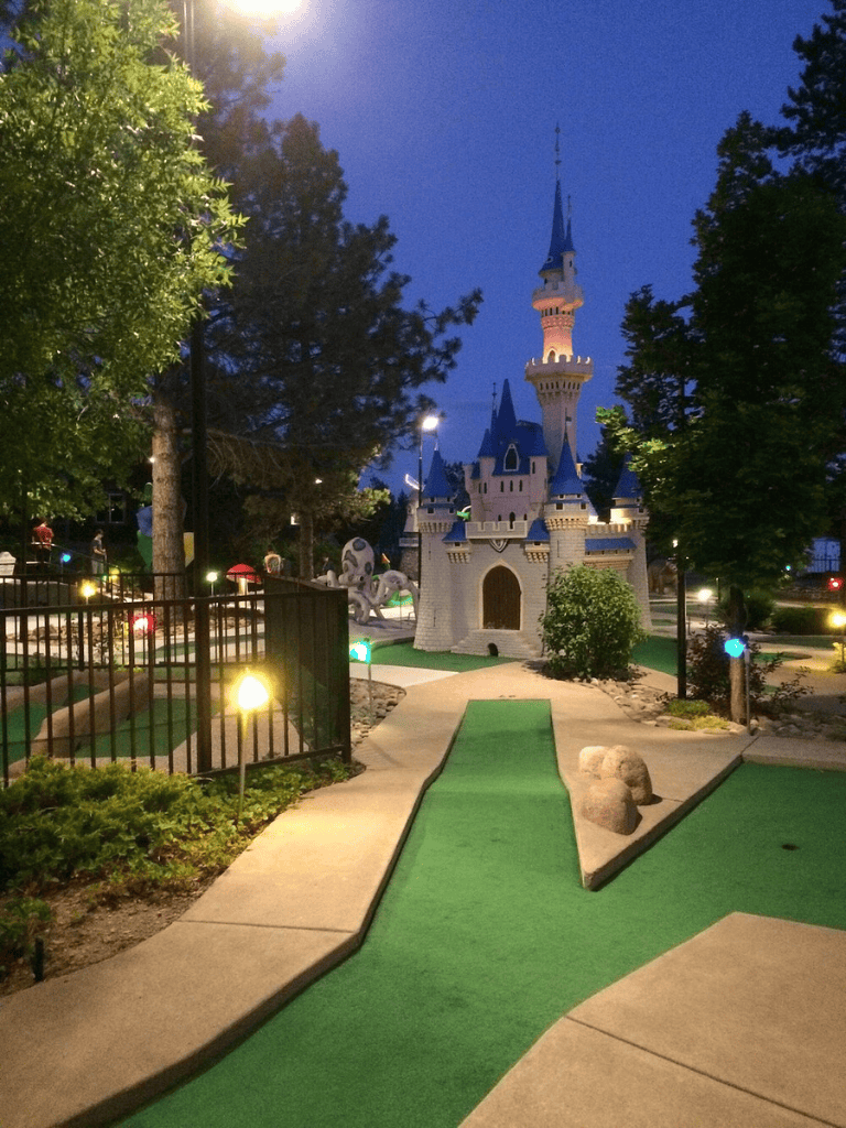 Castle miniature mini golf course at night, illuminated with colorful lights, surrounded by greenery and trees.