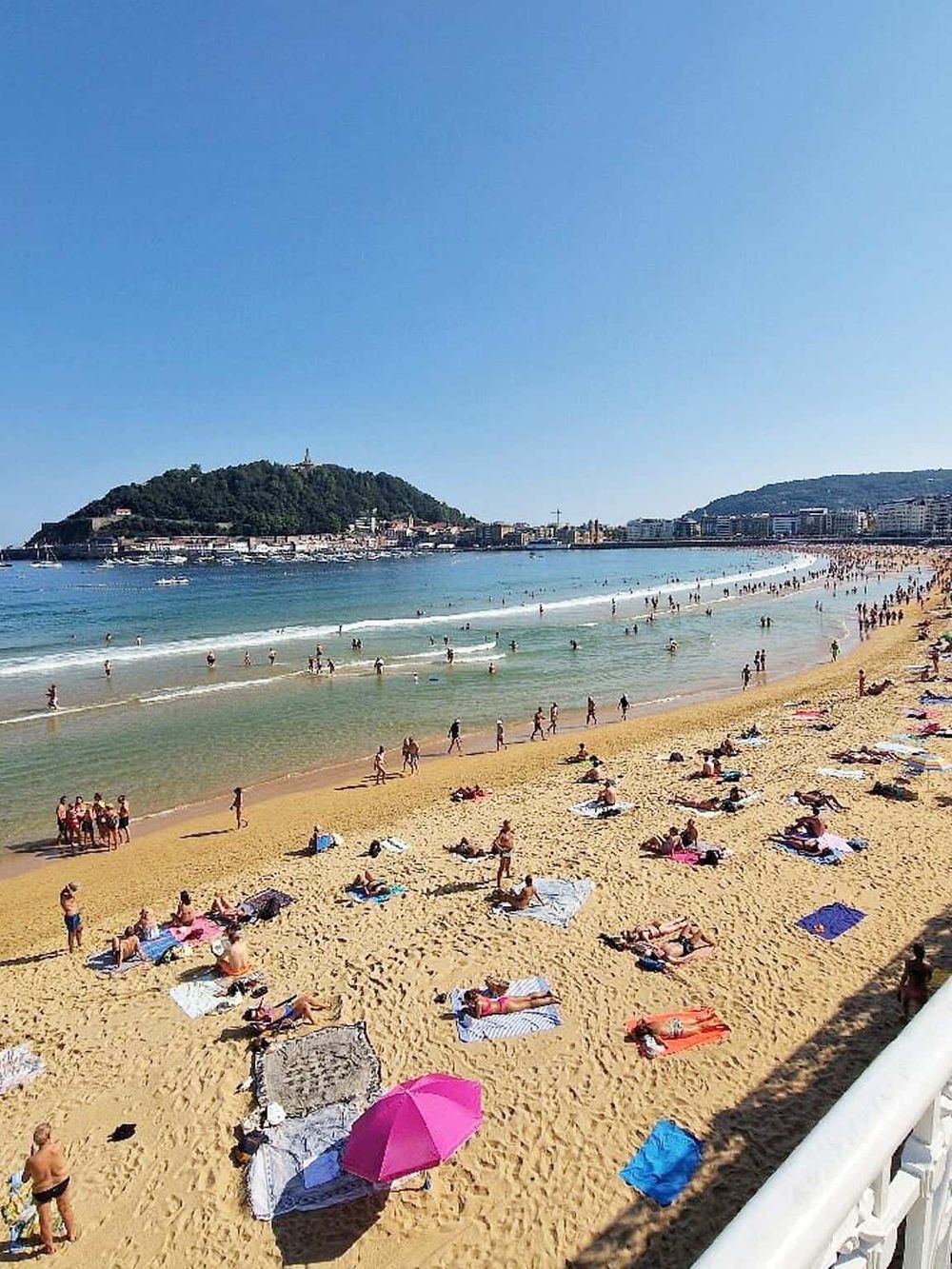 Bustling beach scene with sunbathers, swimmers, and scenic coastal city backdrop in San Sebastián, Spain.