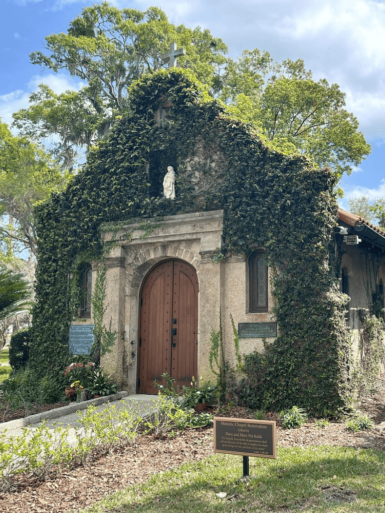 Vine-covered historic chapel with wooden door and religious statues, surrounded by lush greenery.