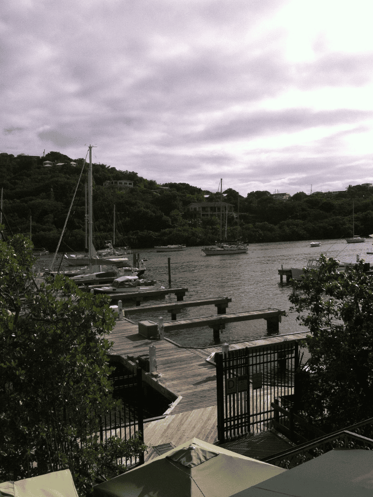 Boats docked at a scenic marina with lush green hills and overcast sky in the background.