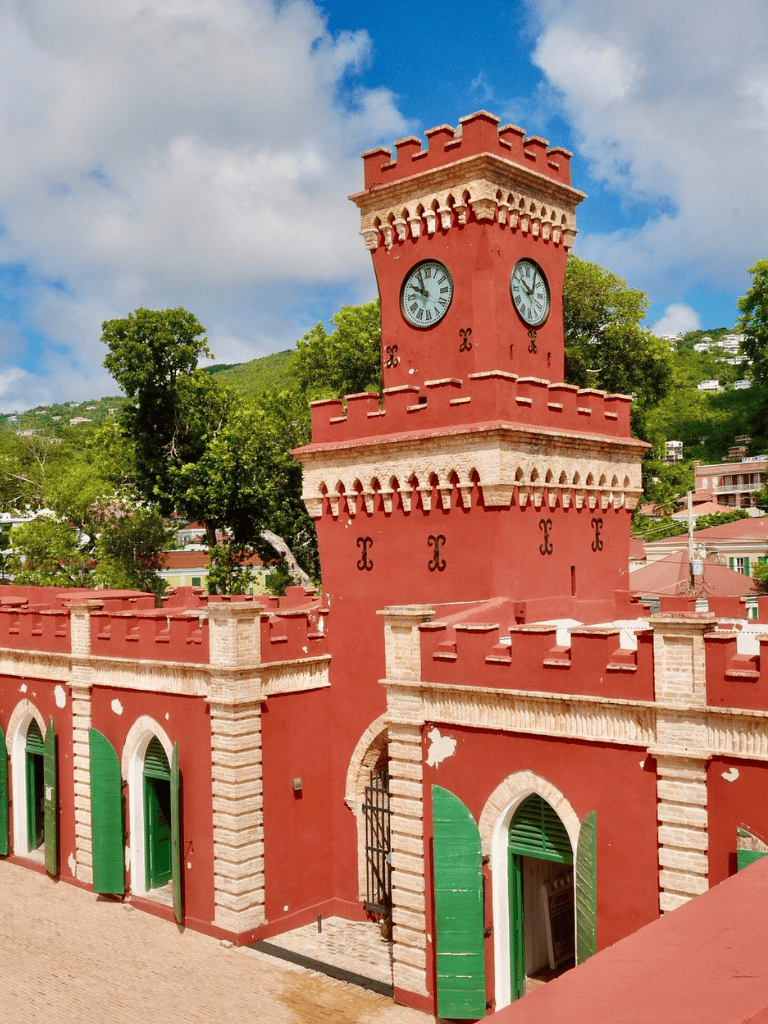 Clock tower with red facade on hillside, historic architecture, vibrant colors, scenic view, tropical climate, local culture, tourist attraction.