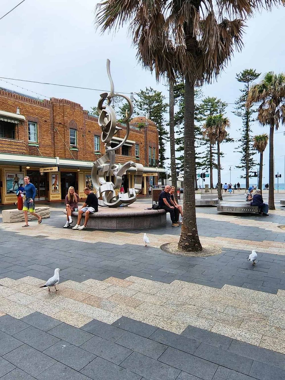 Colorful coastal town street with palm trees, public art sculpture, seagulls, and people enjoying outdoor scenery.