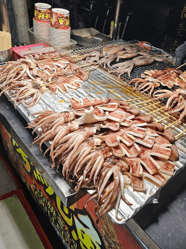 Grilled seafood and meat skewers at a street food stall in Asia, showcasing fresh octopus and sliced pork.