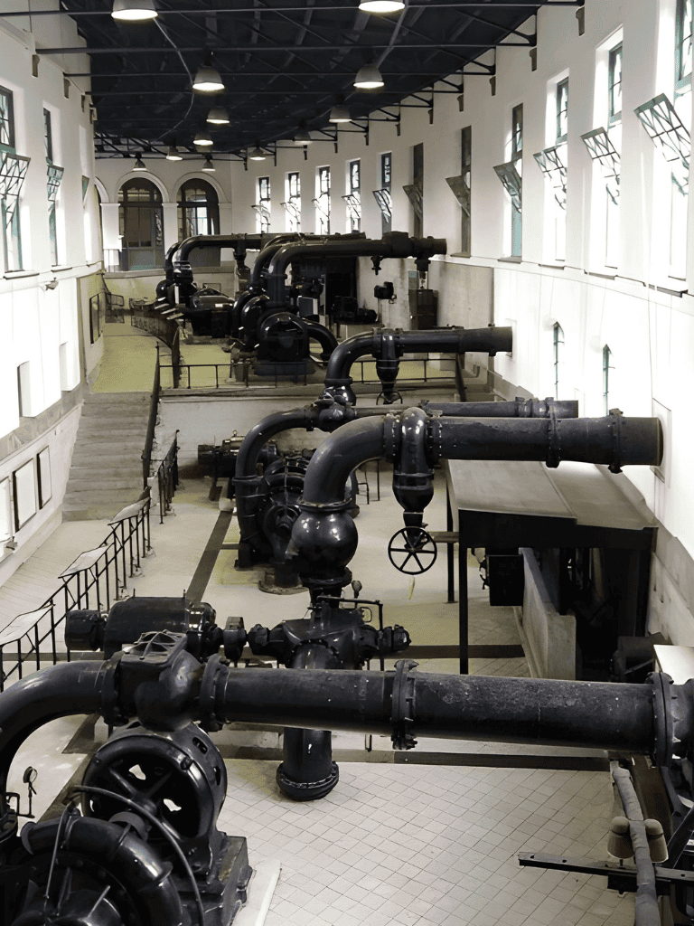 Steel industrial pipes in a historic building with tall arched windows and modern lighting, part of a museum exhibit.