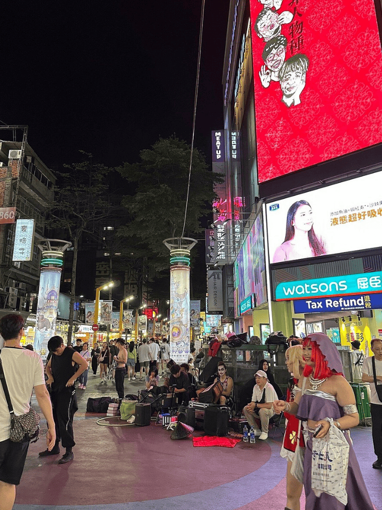 Brightly lit busy city street at night with street performers and digital billboards, showcasing urban nightlife and entertainment.