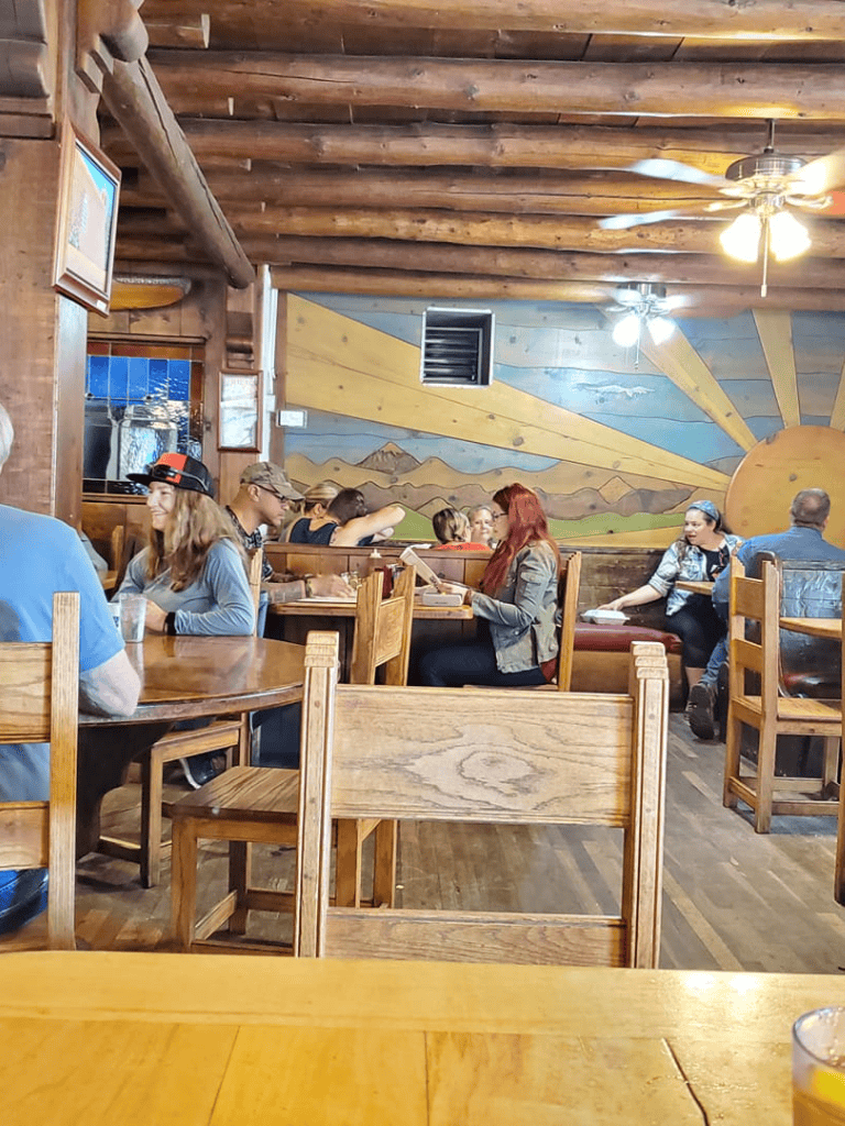 Cozy rustic restaurant interior with wooden beams, mountain mural, and diverse group of people dining.