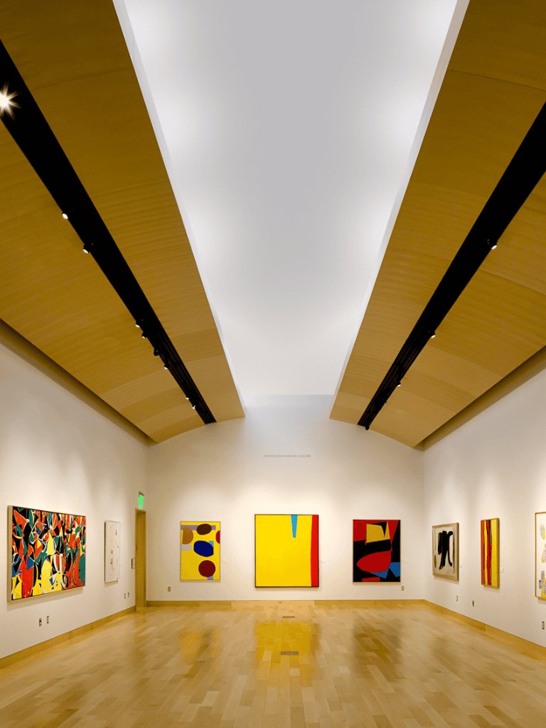 Modern art gallery interior showcasing colorful abstract paintings, wooden ceiling, and hardwood floors.