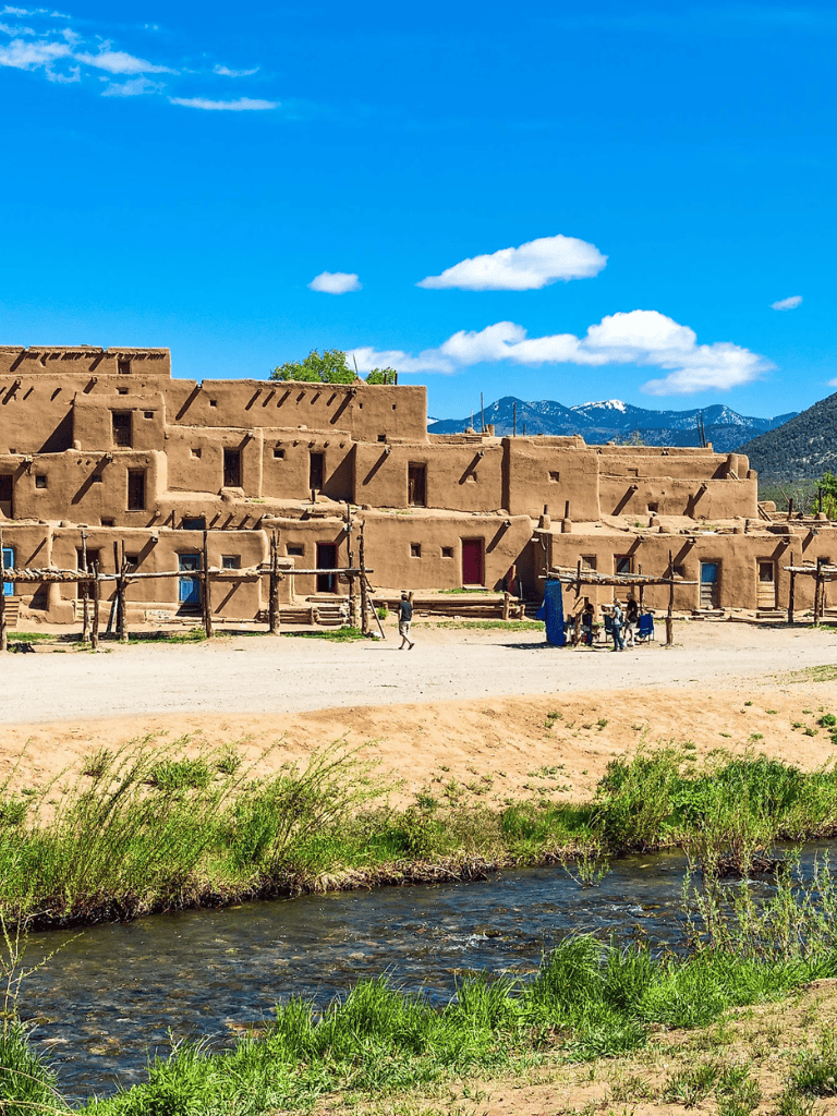 Ancient adobe pueblo with mountain backdrop and flowing river, showcasing traditional architecture and scenic landscape.