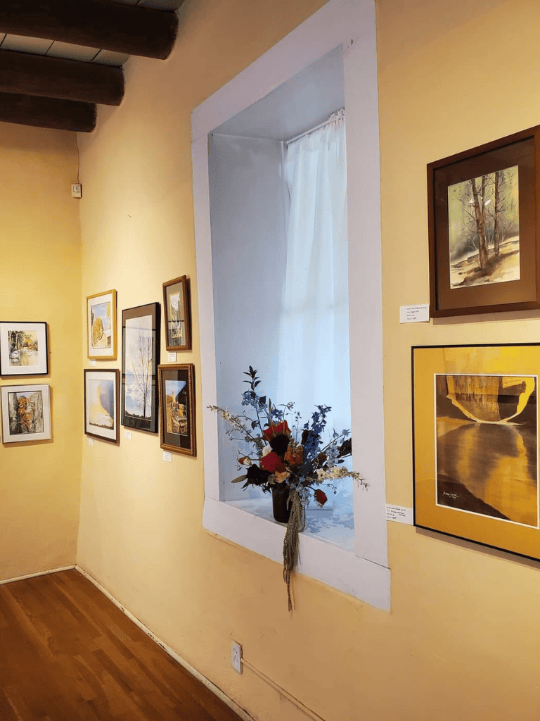 Colorful framed artwork displayed in art gallery with a window and floral arrangement.