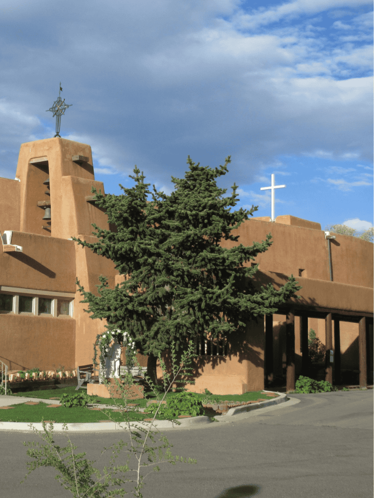 Native American-inspired church with cross, adobe-style architecture, surrounded by greenery and blue sky.