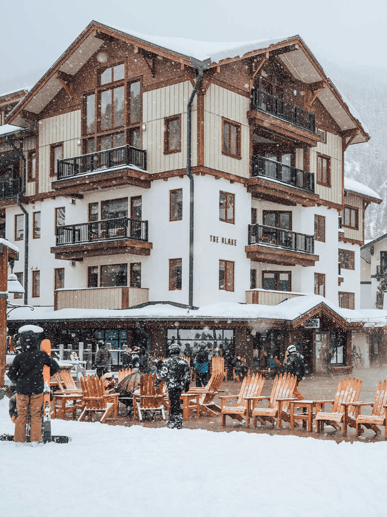 Chalet-style ski lodge surrounded by snow, with skiers and outdoor seating in a winter resort setting.