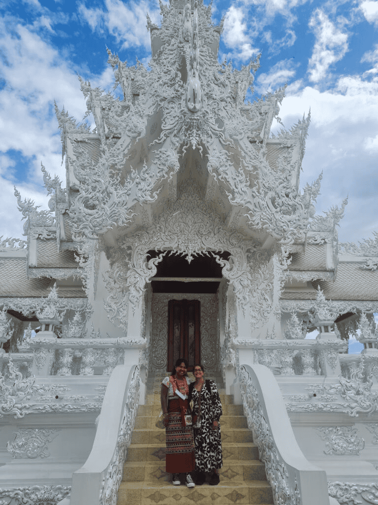 Ornate white temple with intricate carvings and two women standing on stairs under a cloudy sky.