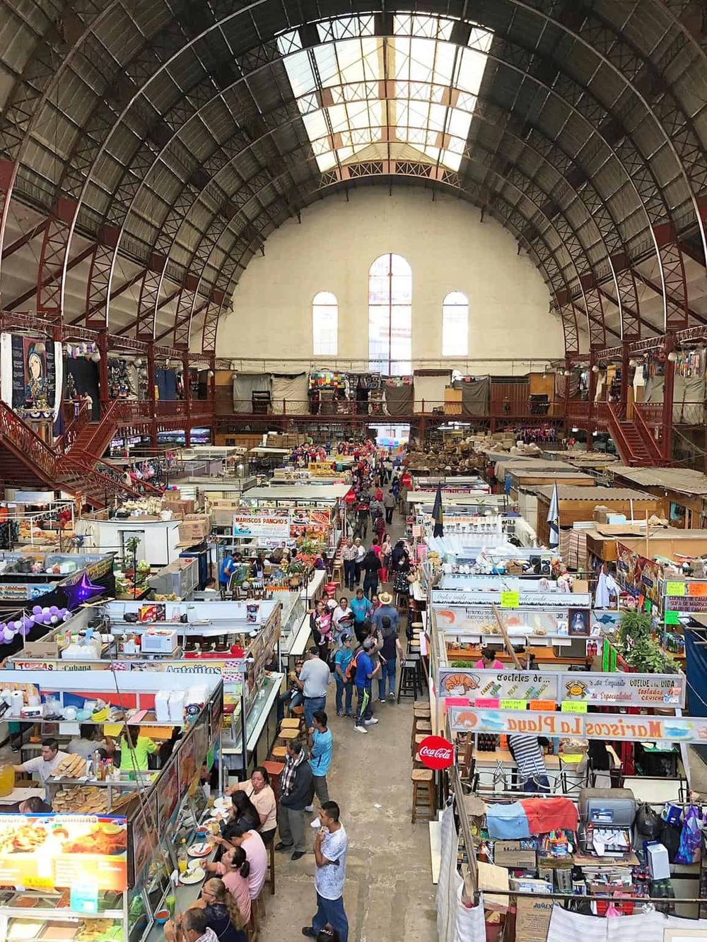 Colorful indoor market with food stalls and shoppers under a large arched ceiling.