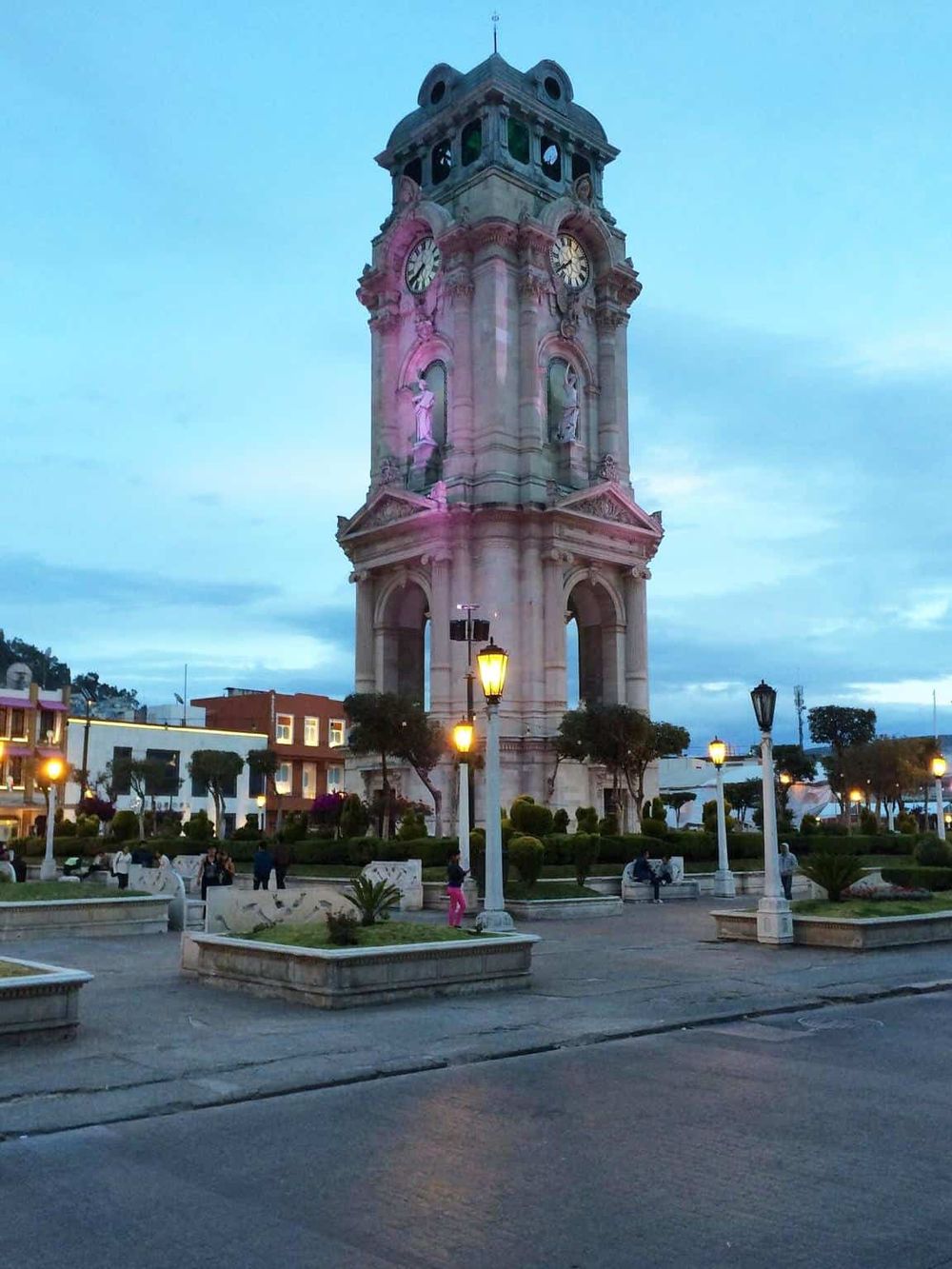 Historic clock tower in downtown, luminous at dusk, surrounded by shops and pedestrian plaza.