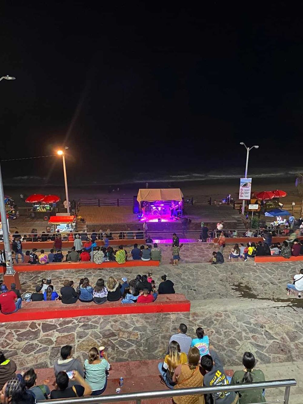 Night beach concert with audience, stage, and ocean view, outdoor entertainment at seaside promenade.