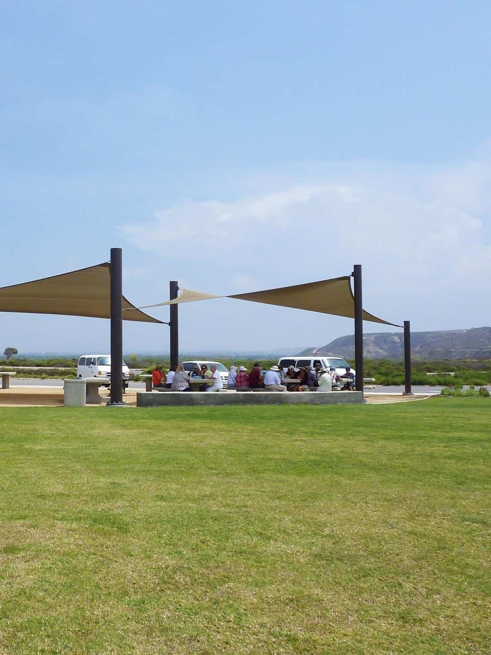 People enjoying outdoor picnic at scenic park with shade sails and vehicles in background.