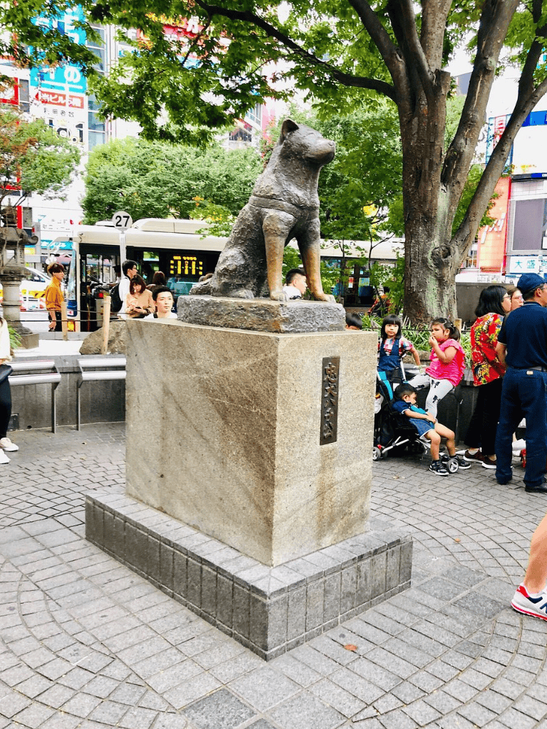 Old dog statue on a city street in front of a busy crowd and trees, reminiscent of Japanese public monuments.