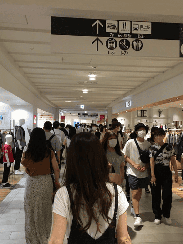 People shopping in a busy mall with directional signs overhead.