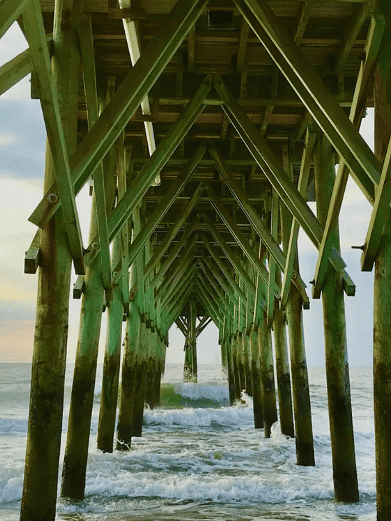Weathered wooden pier extending over the ocean at sunset, showcasing structural beams and seaside scenery.