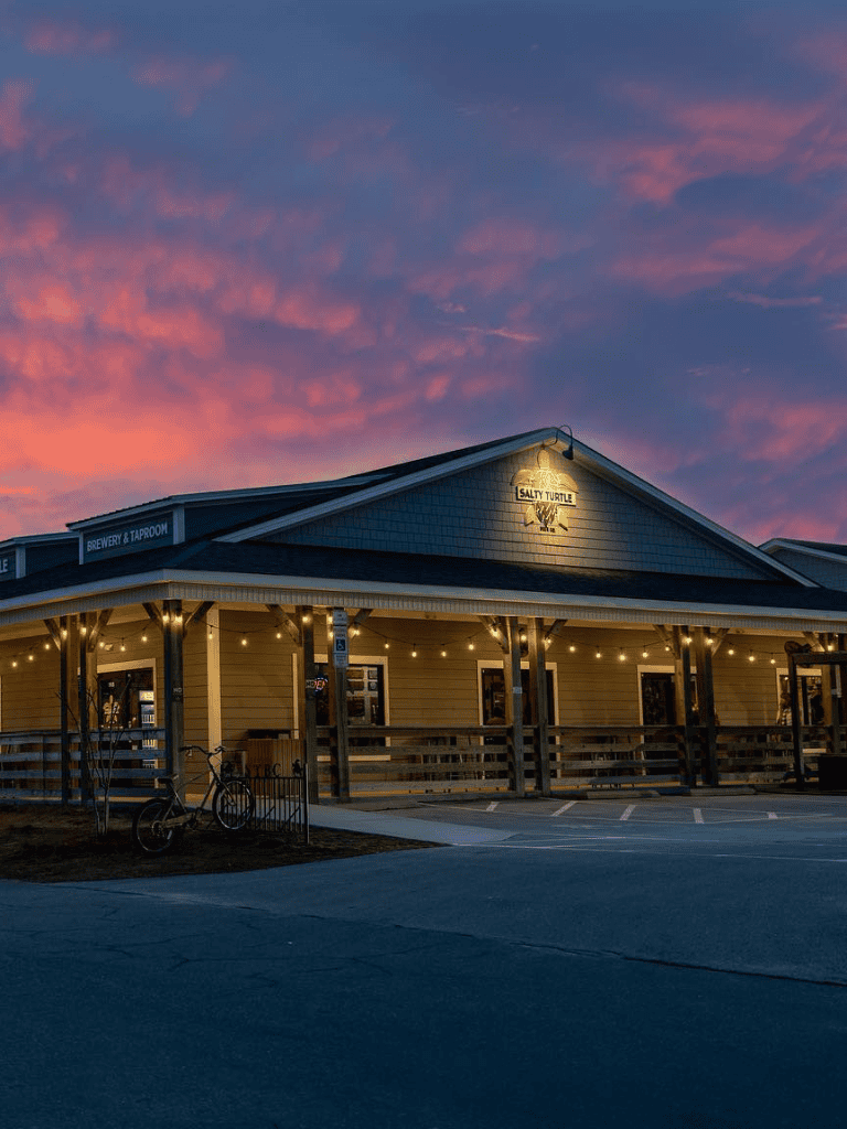 Cozy brewery and taproom at sunset with vibrant pink sky, inviting outdoor seating, and welcoming ambiance for beer enthusiasts.
