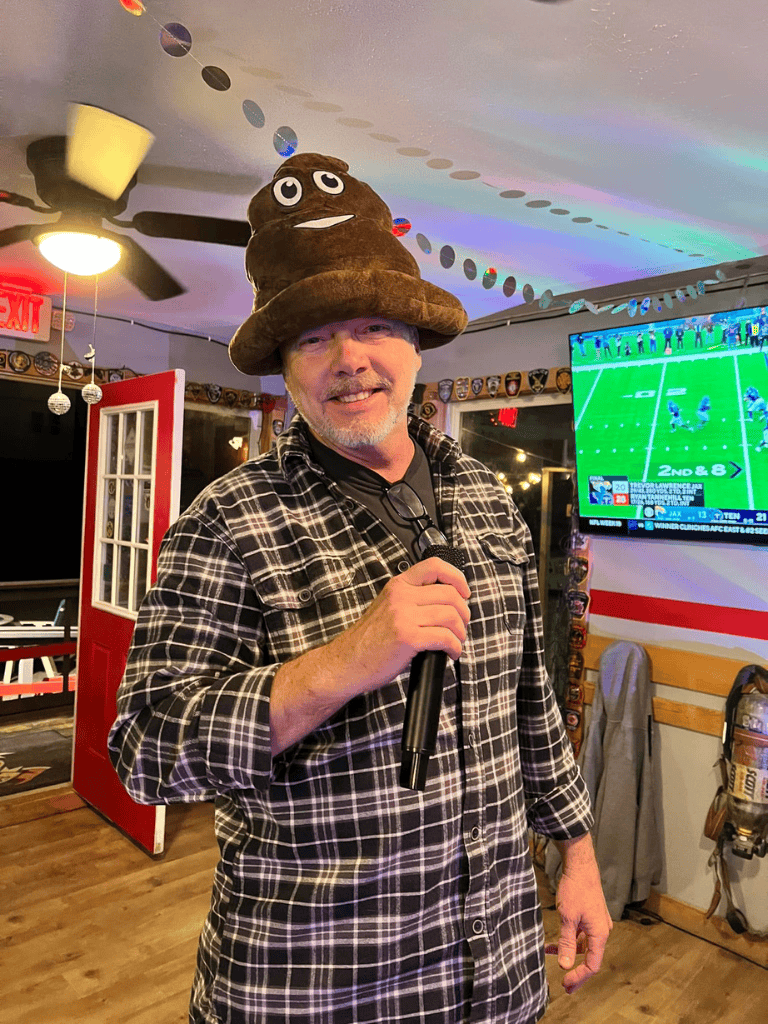 Poop emoji hat, man smiling in plaid shirt, comedy bar, sports bar, QuestForDirections, entertainment, fun pub atmosphere.