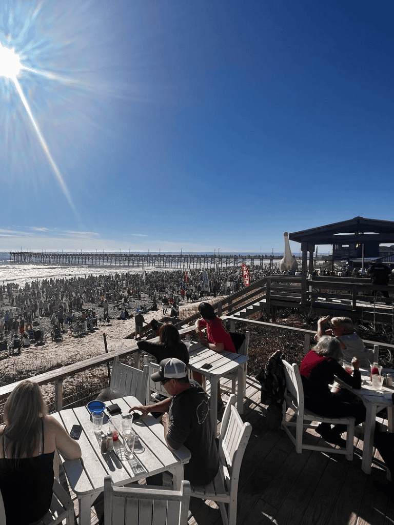 Bustling beachfront restaurant with outdoor seating, ocean view, and sunny sky—perfect for seaside dining.