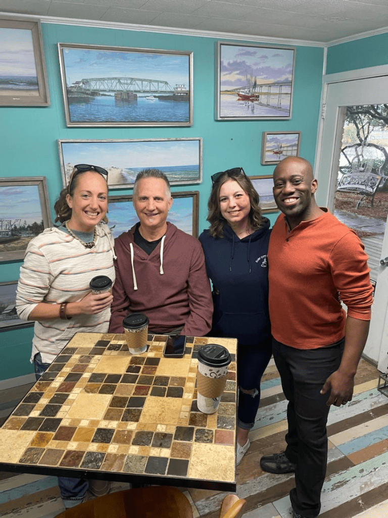 View of four smiling friends in an art gallery surrounded by landscape paintings, enjoying coffee and conversation.