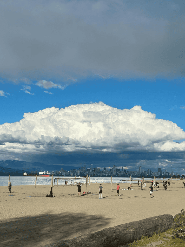 Kids playing volleyball on sandy beach with city skyline and mountain backdrop under cloudy sky.