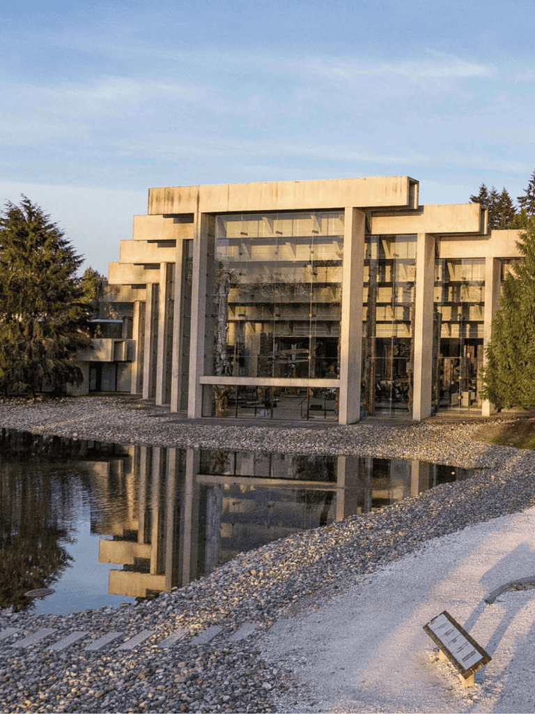 Modern concrete building with glass windows reflected in water, situated near trees and a pebble path.