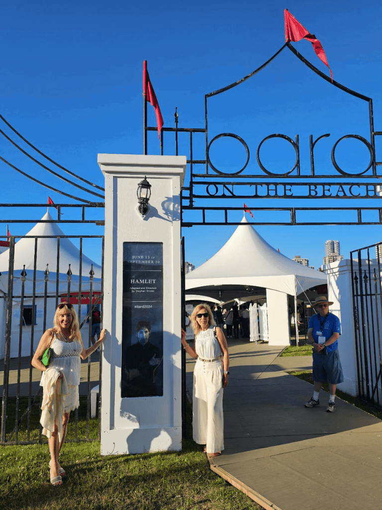 Elegant women at outdoor Shakespeare's Hamlet show entrance, with tents and gates, on a sunny day.