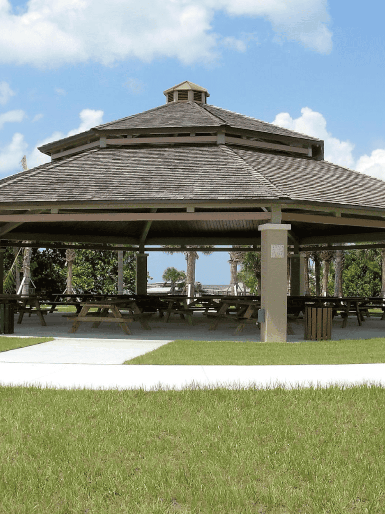 Shaded pavilion with picnic tables near beach and palm trees, ideal for outdoor gatherings.