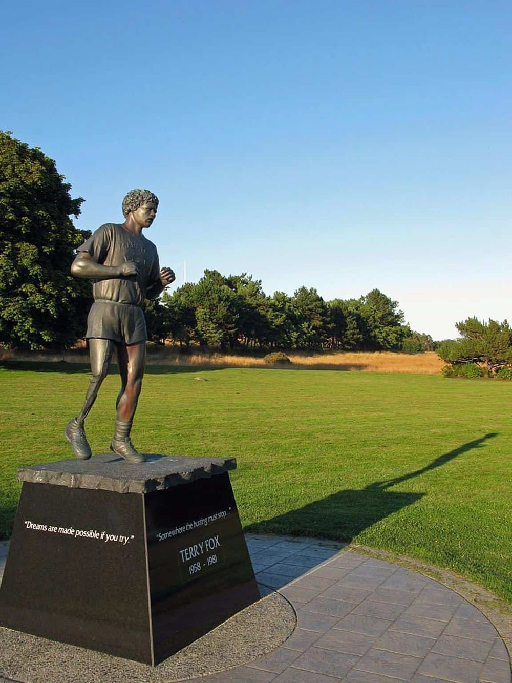 Bronze statue of a runner on a grassy field with trees and clear sky background.